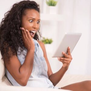 Black woman holding tablet and looking at it in shock.  Holding her face with her right hand.