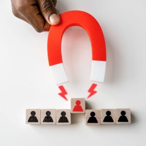 black hand holding a red magnet to signify attracting target audience.  6 people blocks with black figures and one red people block in the center being pulled by the magnet. 