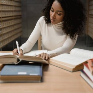 black woman writing at her desk in the library.  Open book  on the side of her.  One closed black book in front of her. 