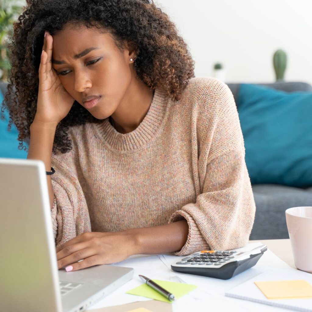 Black woman sitting at her desk in front of her laptop wearing a pink sweater.  Placing her right hand on the top of her head looking frustrated.