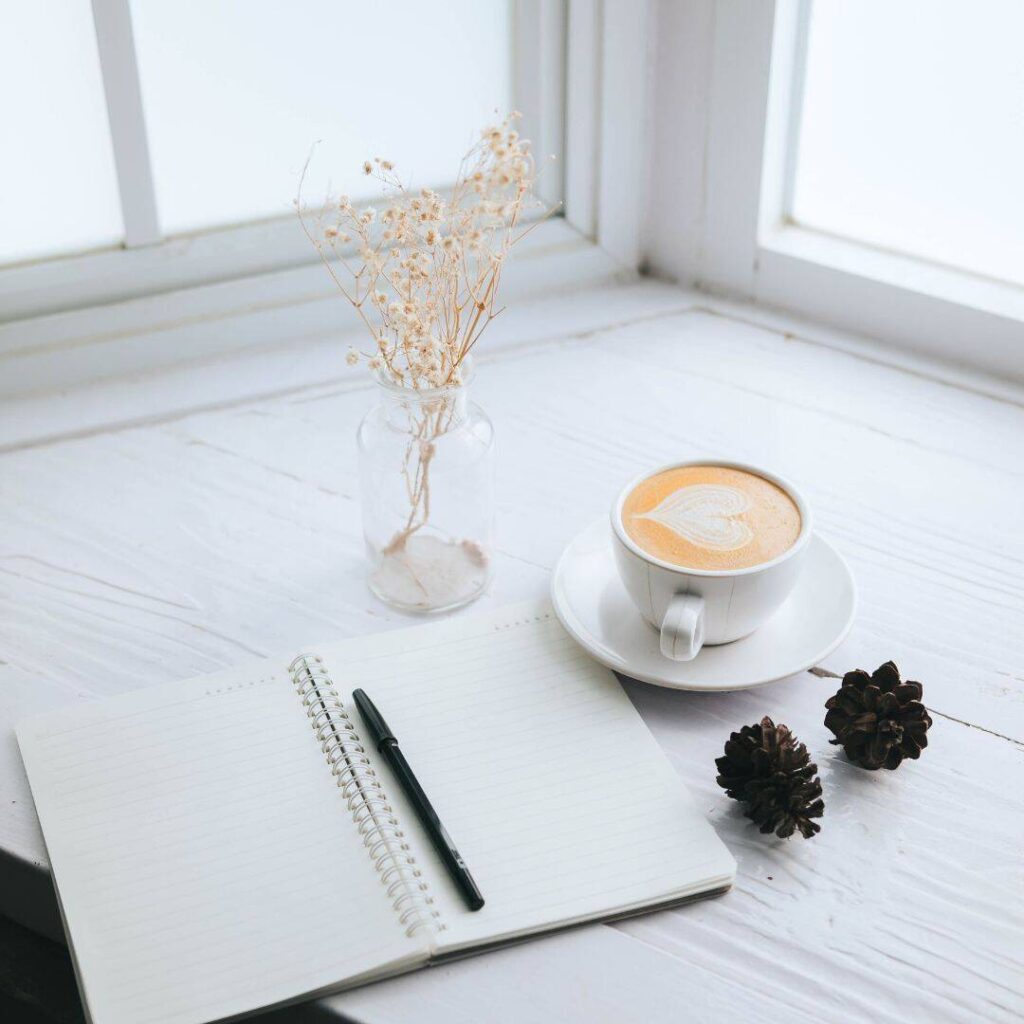 Cappuccino, plant, black pen and notebook sitting on a table by the window.