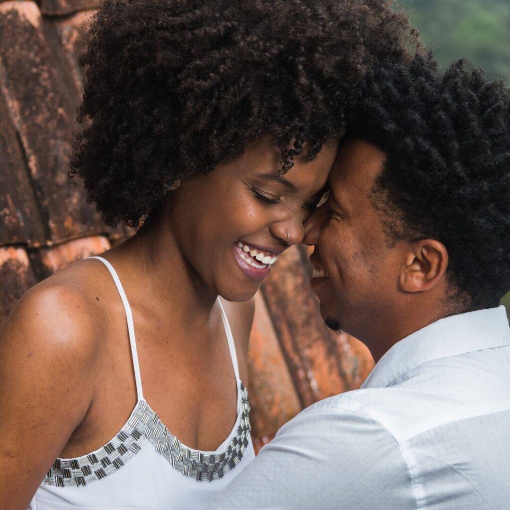 Black couple, man and woman, leaning up against a wall with their heads together smiling intimately.