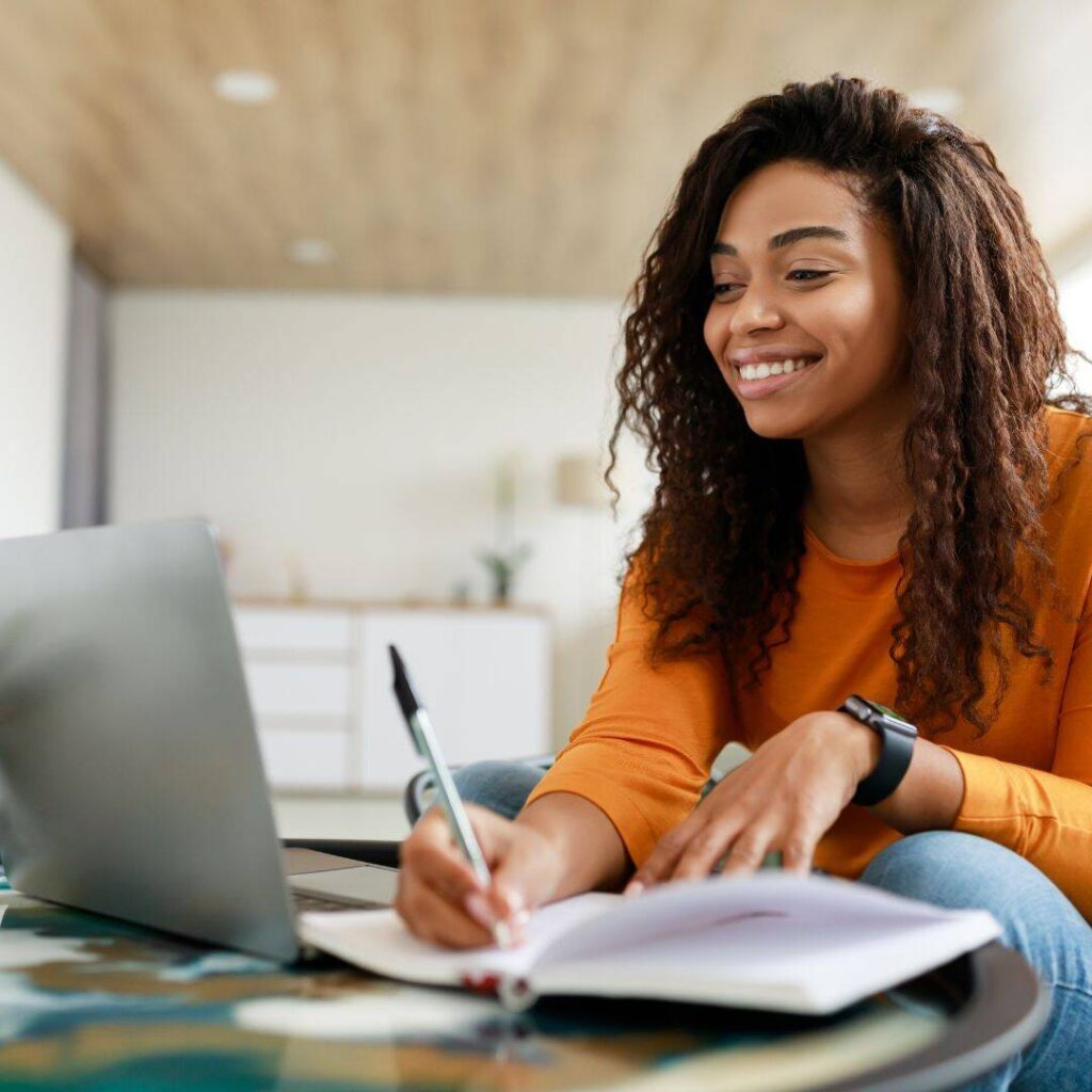 Black woman sitting at a desk writing with a black-capped gray pen and smiling with a gray laptop open in front of her. 