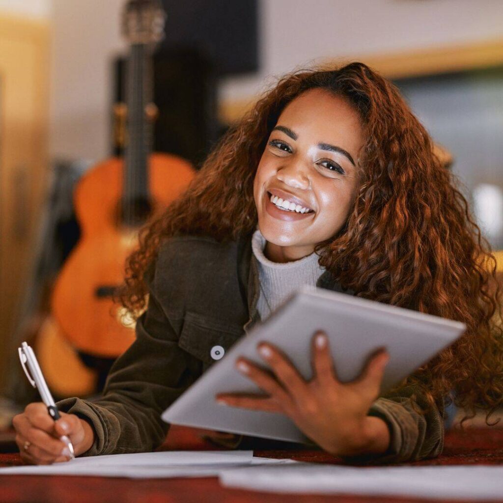 Woman of color at desk holding closed gray laptop in hand close to her chest.  Smiling looking into the camera holding a pen to write.