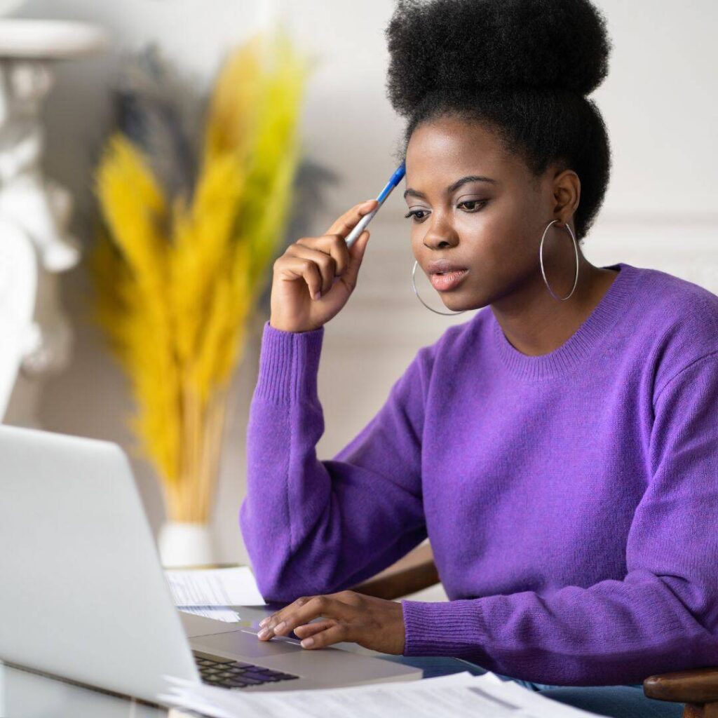  Afro black woman wearing a purple sweater with pen to her head.  Sitting in front of laptop trying to figure out her work. 