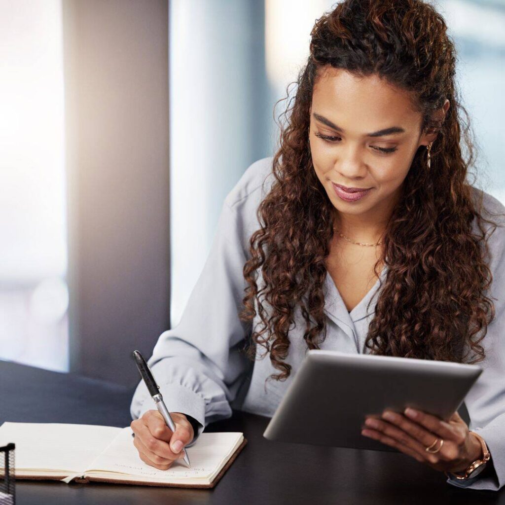  Black woman holding digital tablet slightly smiling while writing in notepad looking at 5 tips to start your book manuscript for aspiring writers. 