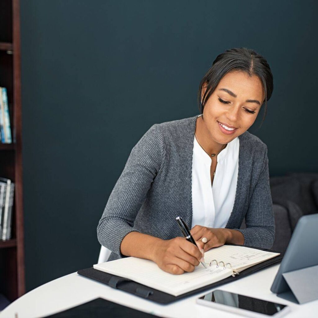  Young Woman sitting at her desk smiling looking down and brainstorming ideas in her book