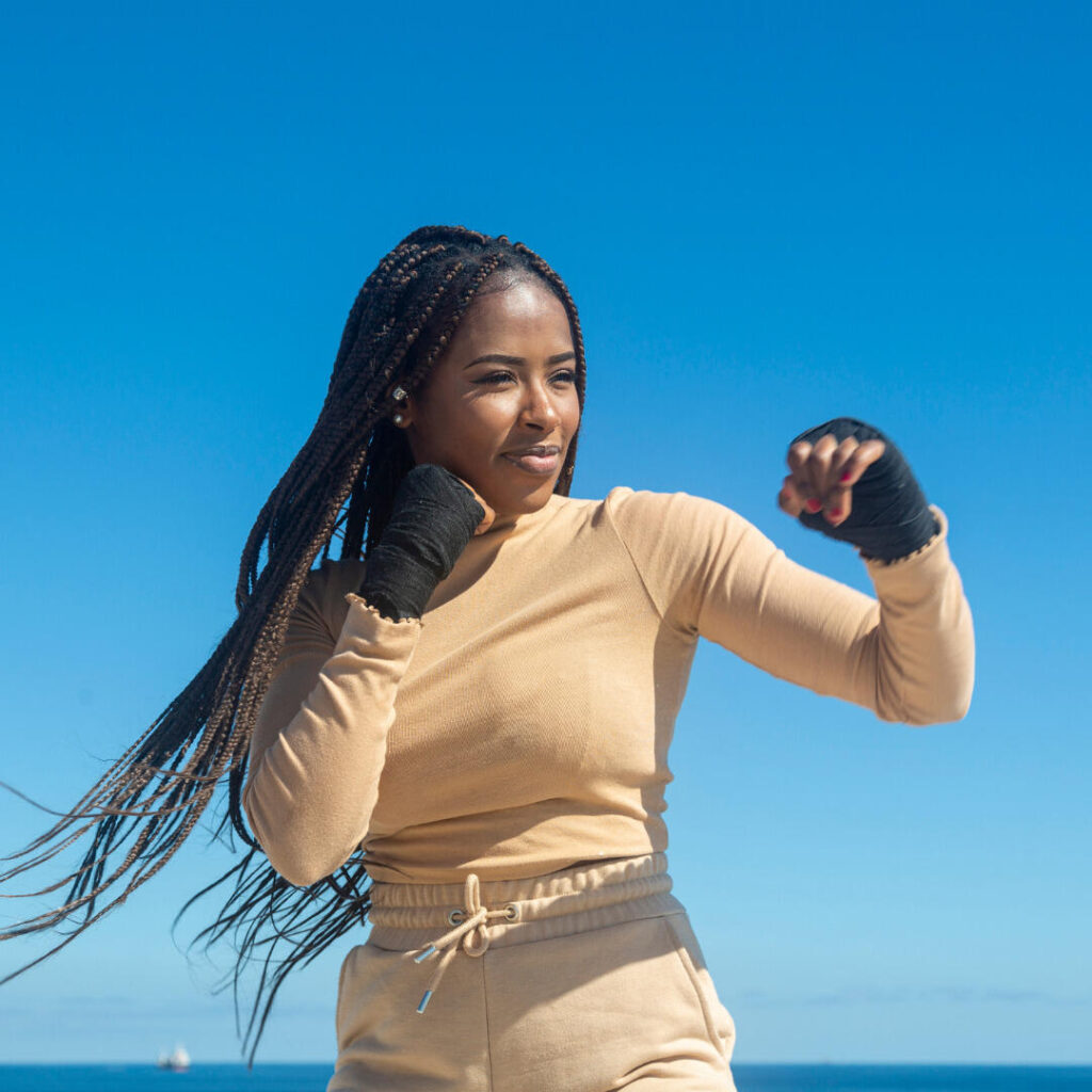 Black woman wearing long braids,  tan turtle neck, tan pants, black hand gloves with open fingers in boxer stance.