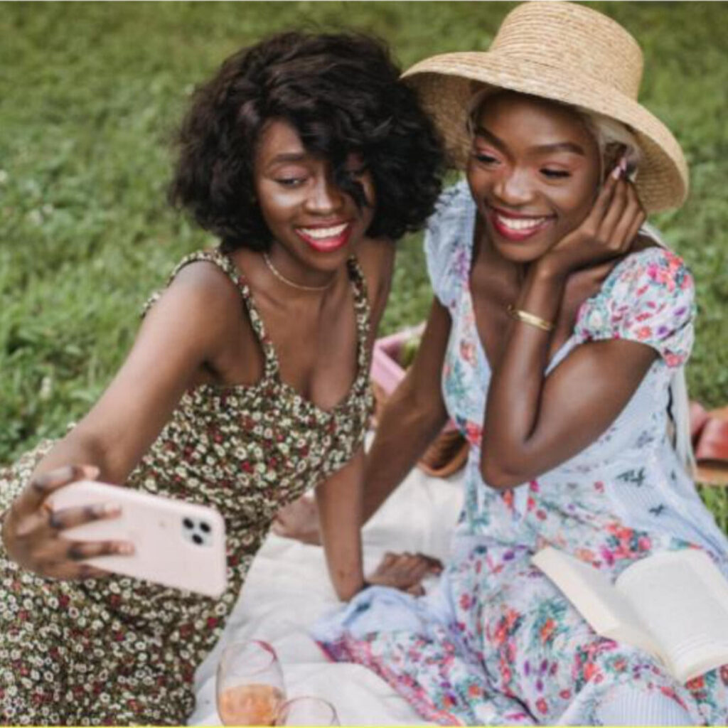 Two black women sitting on a white picnic blanket on the grass. One woman is taking a selfie of them and the other is wearing a sunhat with a book in her lap.