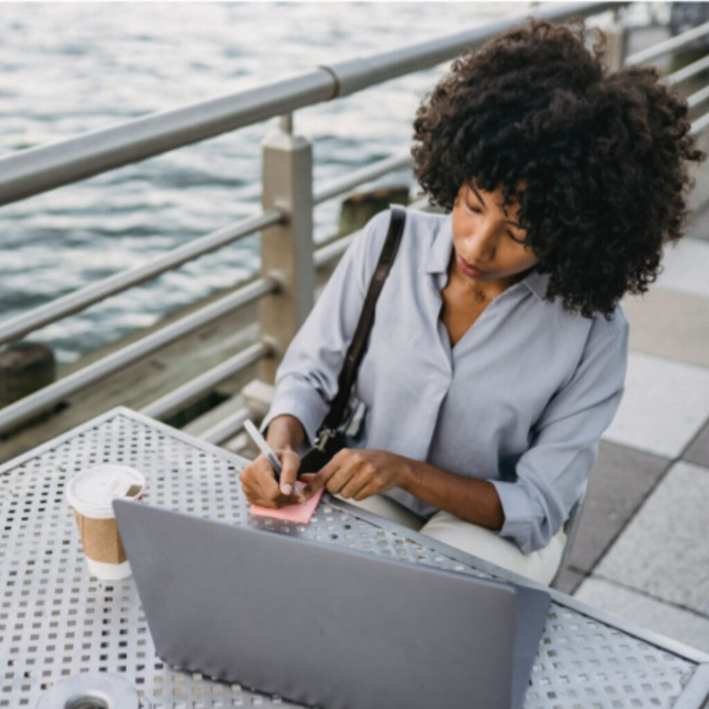 A black woman is sitting at a table next to a waterfront. She is writing on a notepad in front of her laptop.