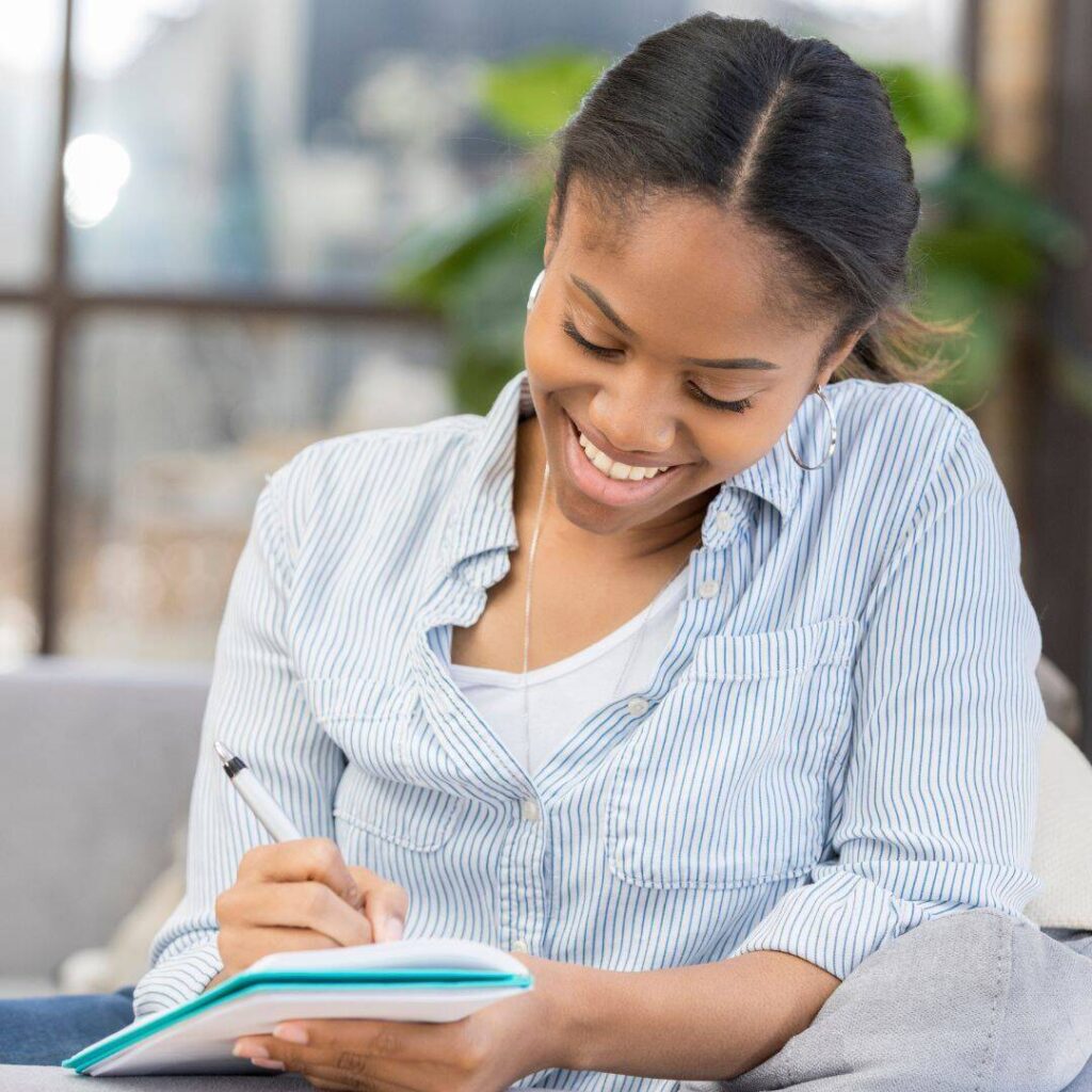 A black woman who is smiling and wearing earbuds as she writes in her notebook.