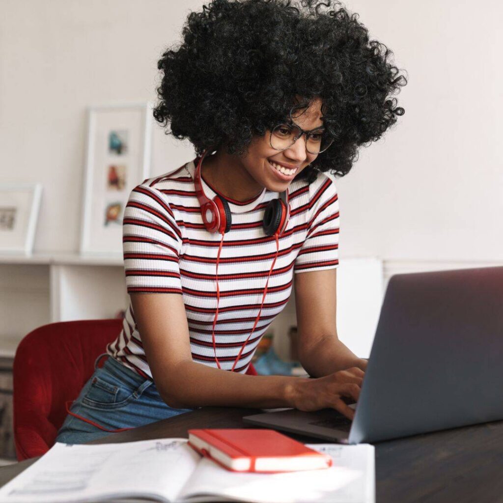 A black woman who is wearing glasses is smiling while typing on her computer. She has red headphones around her neck and a red and white striped t-shirt.