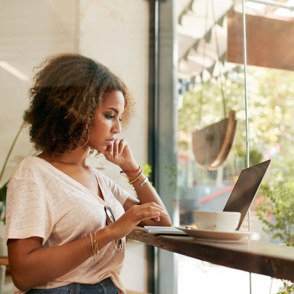 A black woman is sitting at a window counter and typing on her laptop.