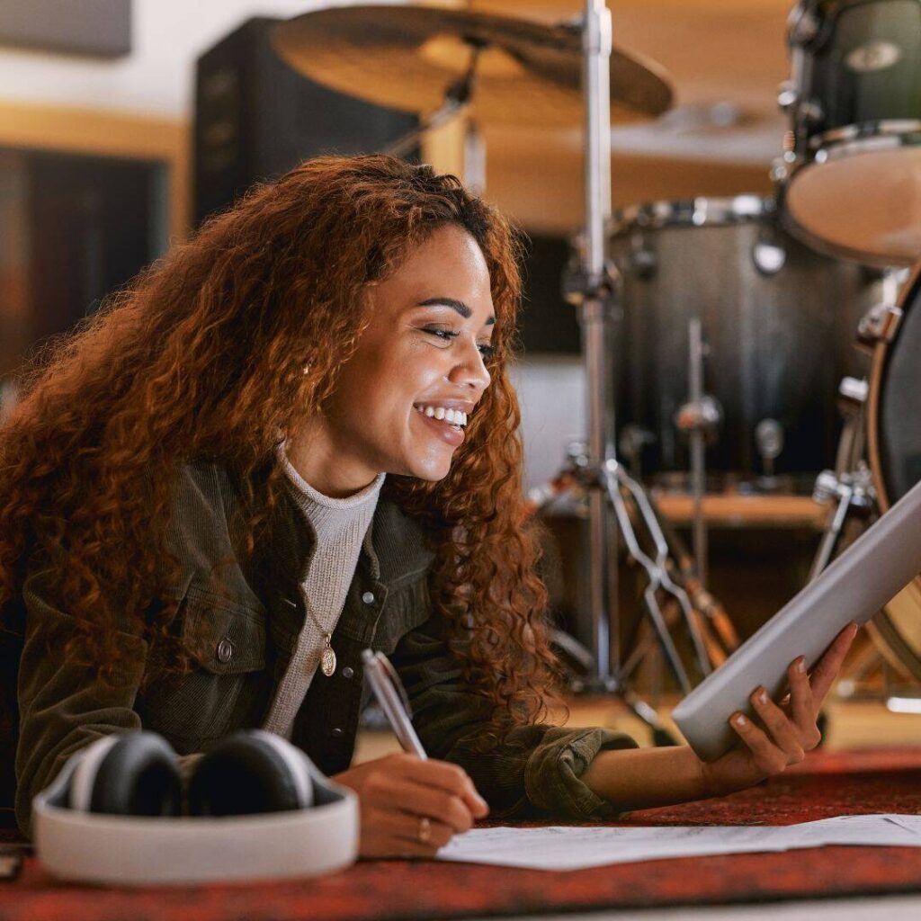 A smiling black woman is on the floor, writing on a paper and looking at her tablet.