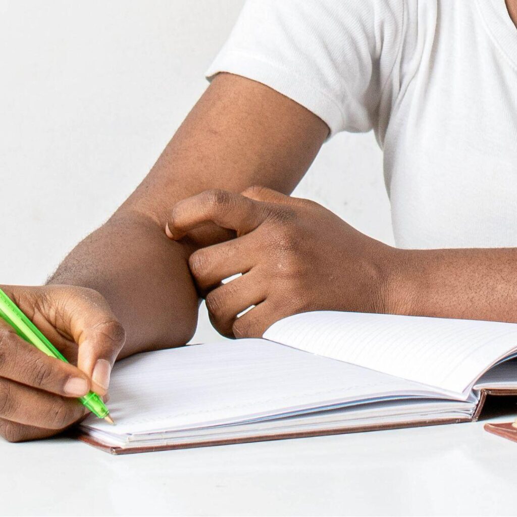 A hand of a black man writing a short story with an open notebook and holding a lime green pen.