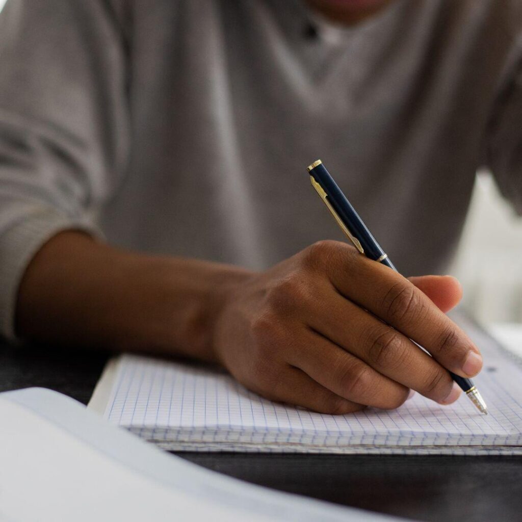 A hand of a Black woman writing on paper with a black and gold pen preparing to write a short story.