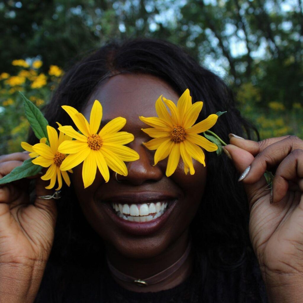 A black woman smiling holding two sunflowers over her eyes.