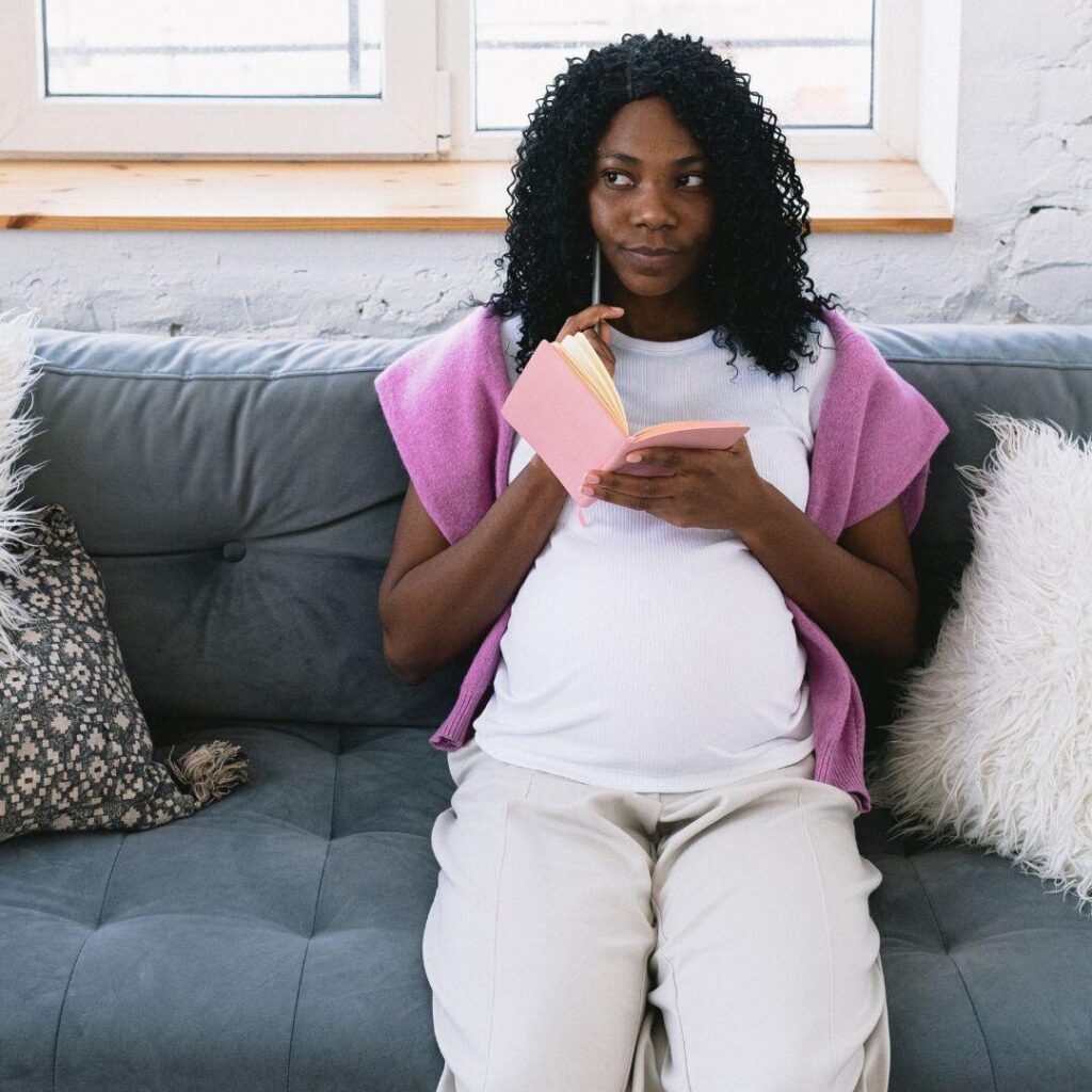 A pregnant Black woman sitting on a teal couch with a fuzzy white pillow beside her thinking with a pin and pink notebook in hand.

