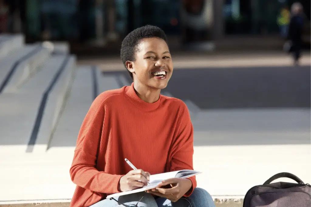 A black woman in an orange shirt is sitting outside and smiling as she writes in a journal.
