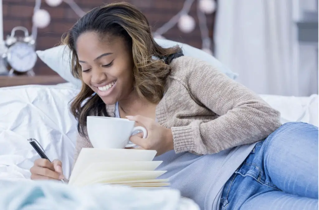 A black woman is relaxing in bed with a white mug in her hand, smiling at her journal as she writes in it with a pen.