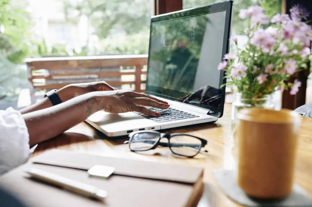A black woman’s hands are typing on a computer keyboard in an outdoor setting. There are a pair of glasses, a vase of flowers, a notebook, and a glass of coffee resting on the table next to the computer.
