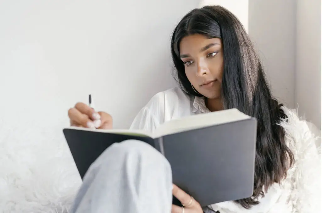 A woman of color is sitting and gazing at an open black journal which she holds in one hand. She has a pen in the other hand and is writing in the journal.