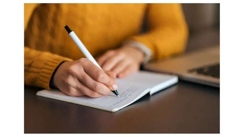 Qualities of a good writer writing with a white, black capped pen in the notebook sitting at a desk wearing a golden yellow sweater.
