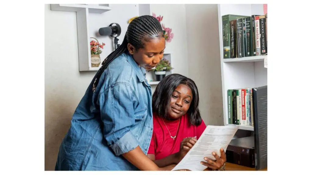 Black woman with braids and denim shirt leaning over her daughter, looking at her paper to help her become a good writer.