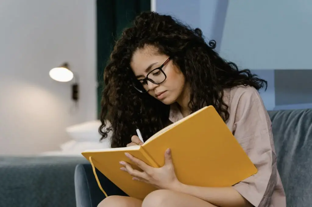A woman of color is sitting indoors and writing in a big yellow journal. She is wearing glasses and seems to be very focused.