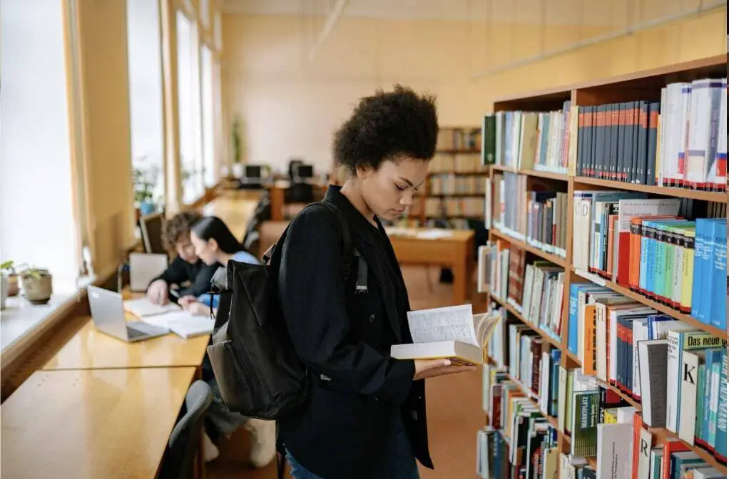 A black woman is standing in a library next to a shelf of books. She is wearing a black
backpack and looking at a book in her hand.