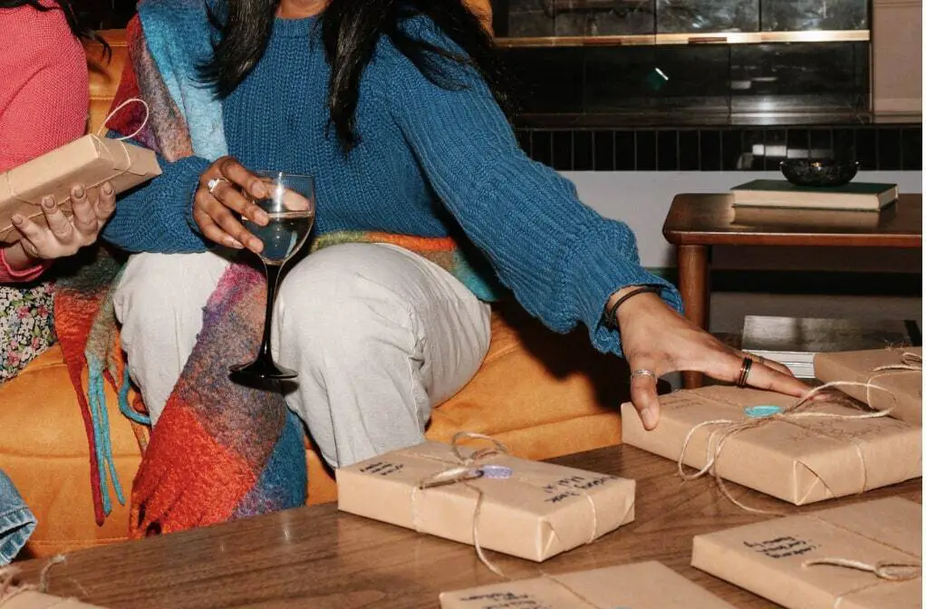 Two women in vibrant colored clothing are sitting on a couch. In front of them is a
coffee table with a few rectangular gifts wrapped in brown wrapping paper. One of the women is
holding a glass of water and touching a gift on the table, and the other woman has a gift in her
hand.