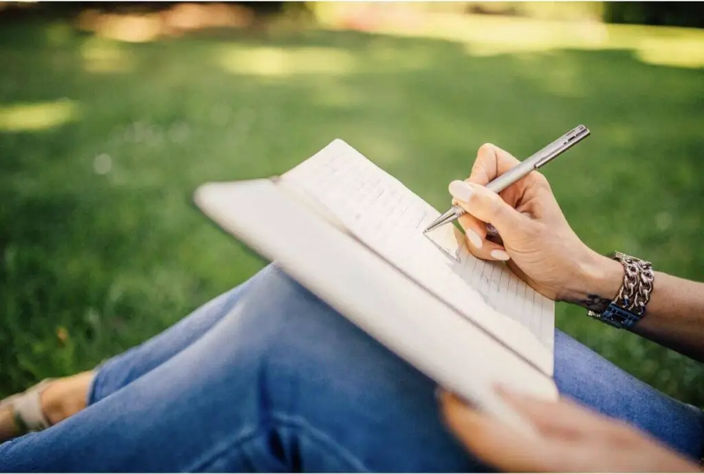 The hand of a woman is shown writing in an open journal. The journal is resting on the woman’s legs, and she is sitting outside in the grass.