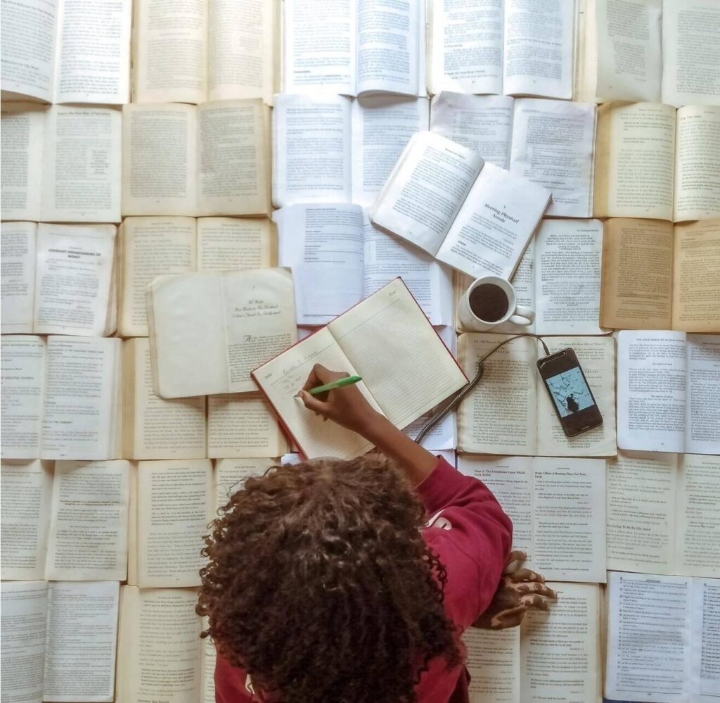 An aerial view of a black woman lying on a floor surface made of open books. She is
writing in one of the books closest to her, and a phone and a coffee mug are placed nearby.