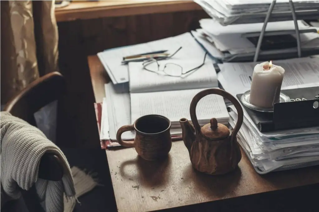Various stacks of papers, a tea kettle, a mug, a candle, a book, and a pair of glasses
are lying on a wooden desk. A wooden chair with a white sweater draped over it is set in front of
the desk.