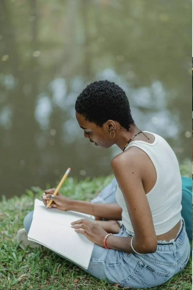 A young black woman is sitting on the grass by a lake, writing in a journal with a pencil.