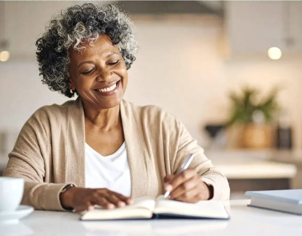 A seemingly-older Black woman is sitting at a table in a kitchen and writing in a journal. She is smiling as she writes.