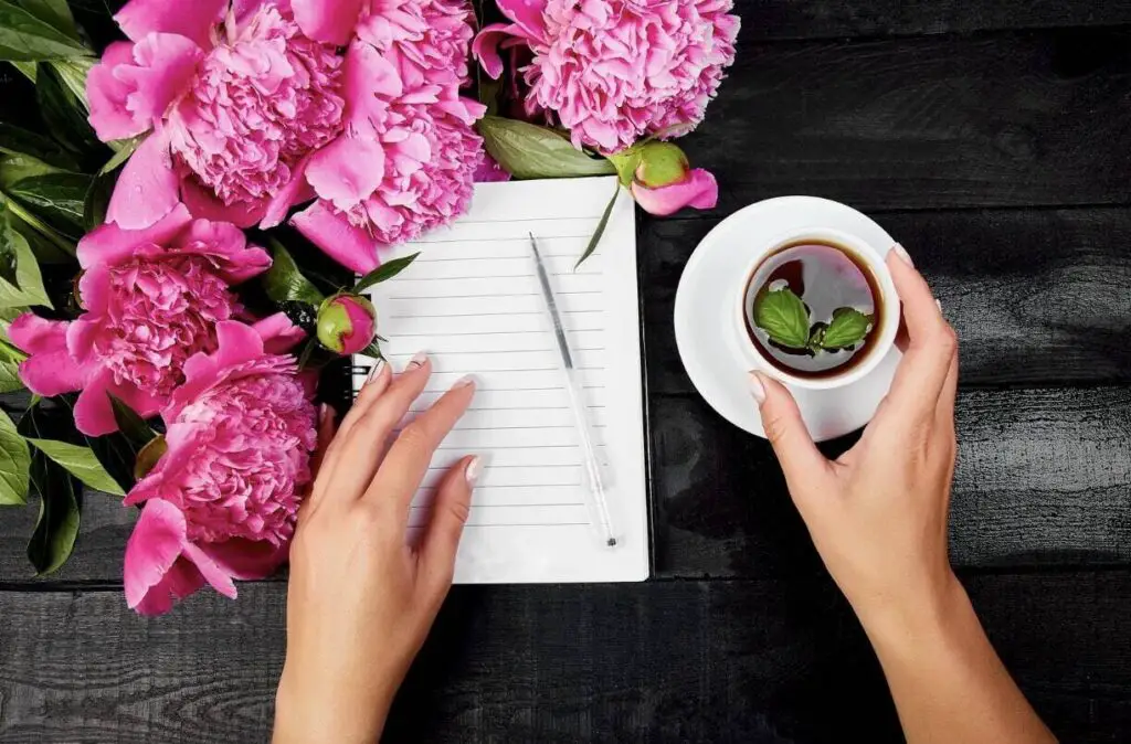 On a dark wooden table, a journal lies open to a blank page. A pen lies on top of it, and next to this setup are numerous pink flowers. On the other side of the journal is a cup of tea on a saucer. There is a woman’s hand resting on the journal, and her other hand is touching the cup of tea.