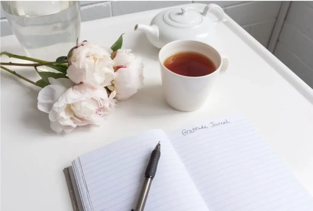 A journal lies open to a page in which the words “Gratitude Journal” are written at the top. A small, pink bouquet of flowers and a kettle and cup of tea lie next to the journal. On the journal itself rests a pen.