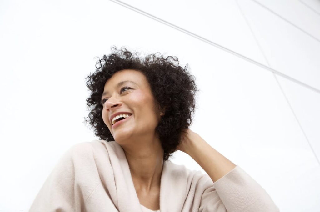 A Black woman is shown with a big, genuine smile on her face. She is looking into the distance and touching the back of her neck with one hand.