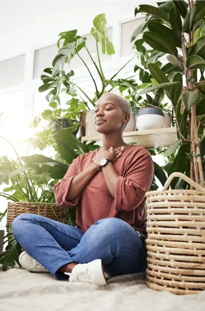 A young Black woman is sitting and meditating. She is surrounded by plants, and her arms are folded across her chest. There is a serene expression on her face.