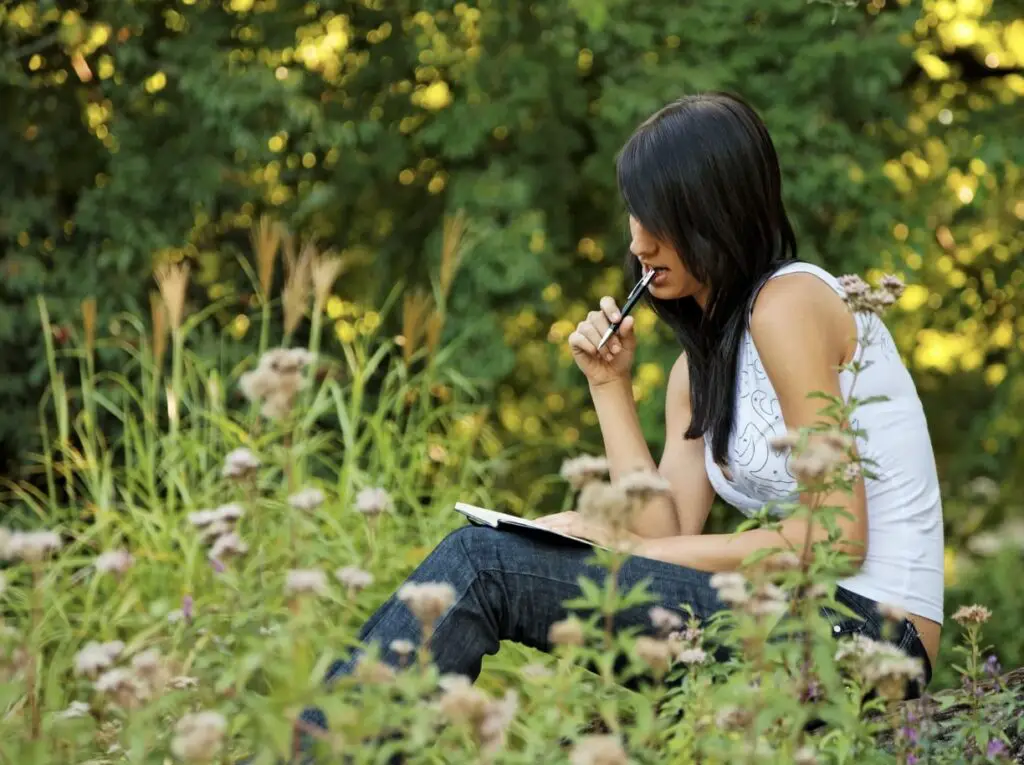 A woman is sitting outside in a meadow-like setting. There is a journal in her lap, and she is holding a pen against her lips with a pensive expression on her face.