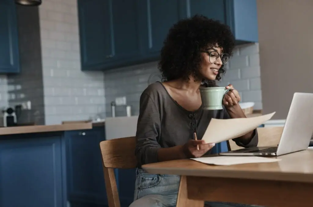 A young Black woman is sitting at a wooden dining table She is looking over various paper pages and holding up a mug to her lips. She has a pleasant expression on her face.