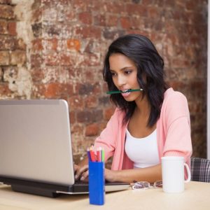 black woman wearing pink hoodie typing in front of laptop with pen in her mouth and white mug sitting beside her. 