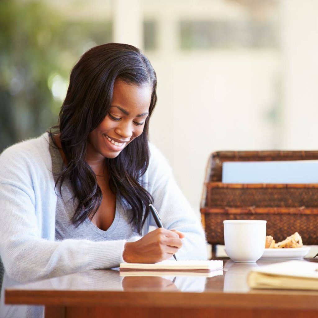 Black woman smiling while sitting at her desk and writing in her journal.