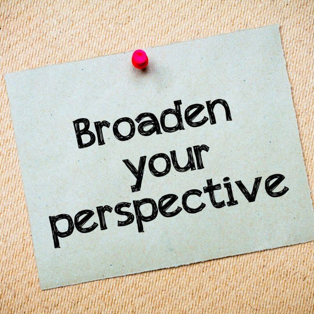 Broaden Your Perspective written on a green square of paper and pinned to a wall with a red thumbtack.