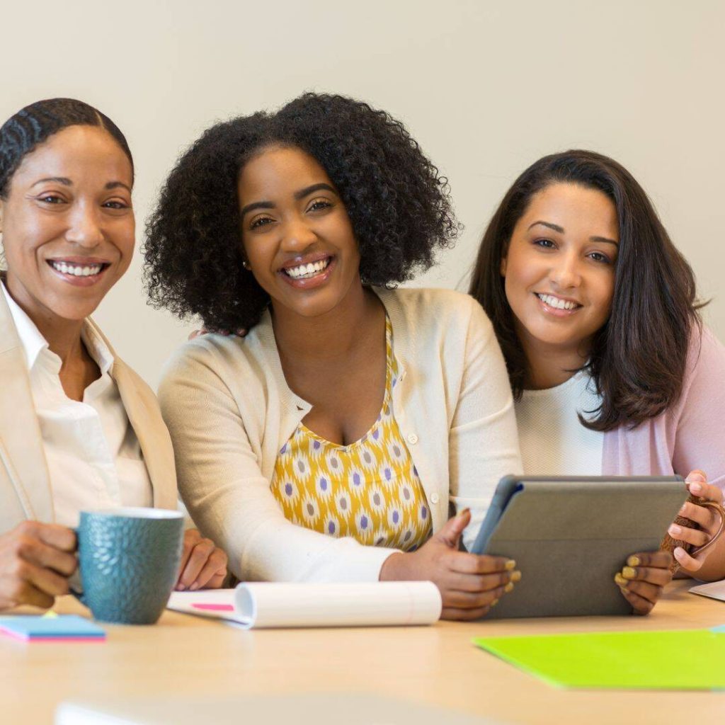 Three Black women of color smiling.  One holding a tel mug, and the other two holding a notebook.  Practicing commmunication skills. 