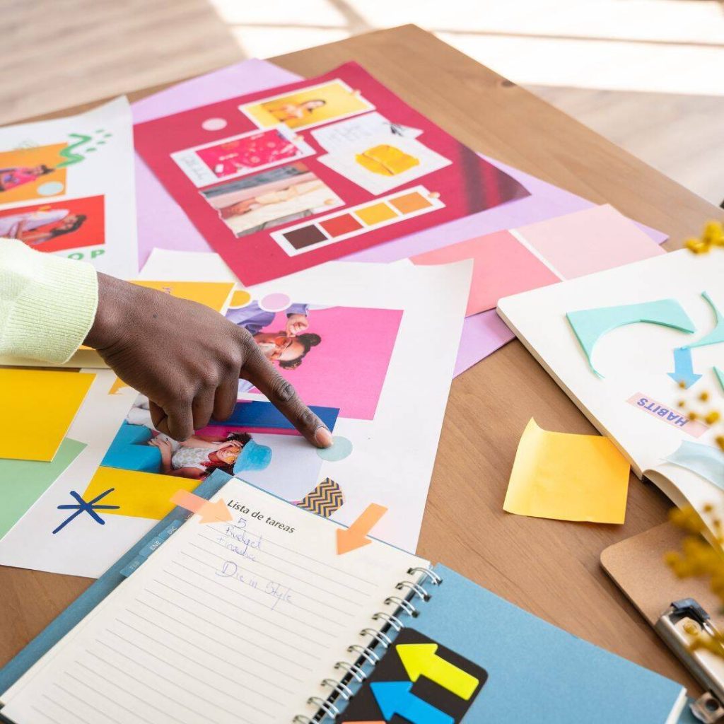 Images, construction paper, and notebook laid out on a table in a creative way.  Black female hand pointing to a blue dot on the paper.