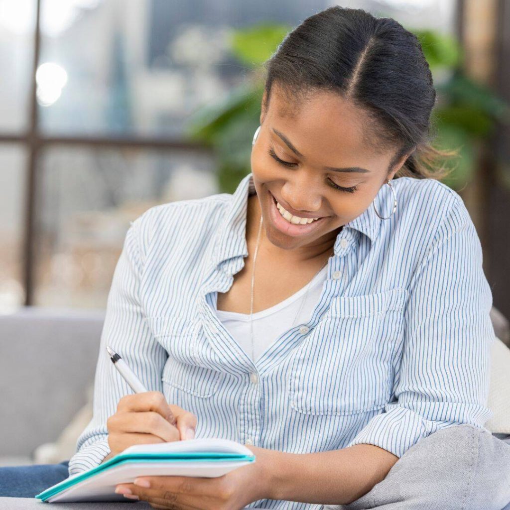 Black woman smiling looking down writing in her notebook.