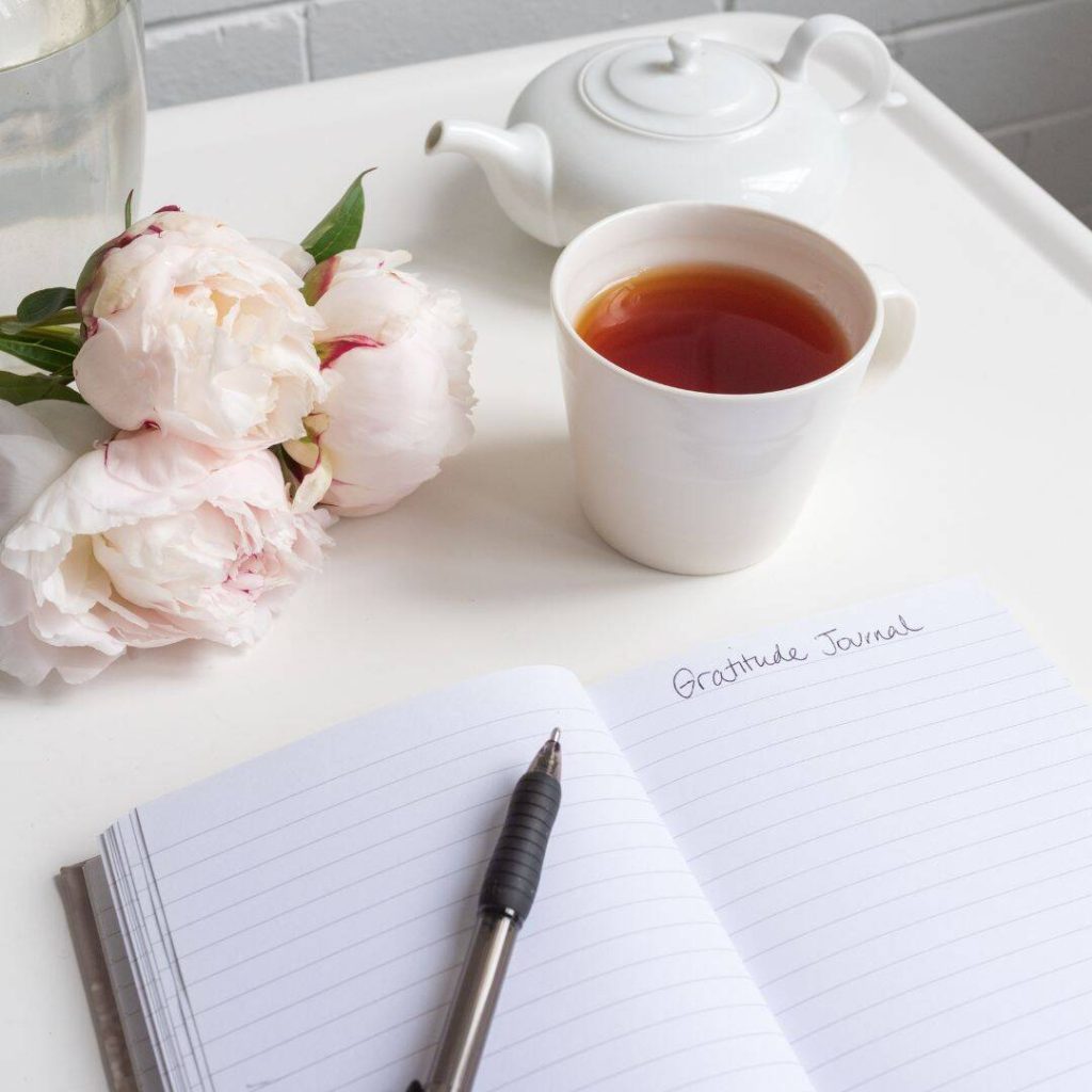Gratitude Journal written in notebook with light pink flowers beside notebook.  On white table with tea cup. 