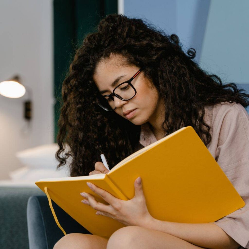 Woman of color with glasses sitting down holding a golden yellow notebook writing in it.